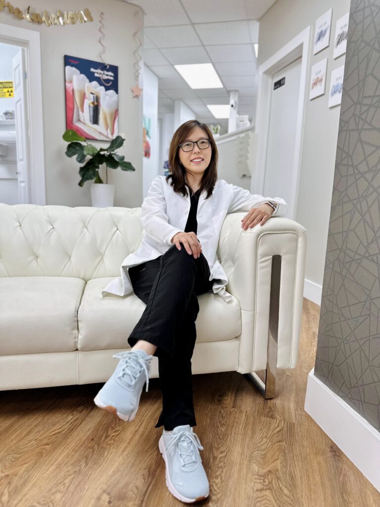 A woman in a white lab coat sits on a white couch in a modern office setting, smiling at the camera.