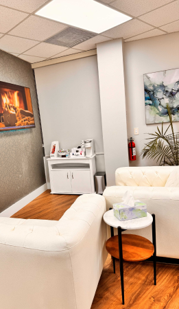 A small waiting room with two white sofas, a round side table with tissues, a cabinet with a coffee maker, a fire extinguisher, and paintings on the walls.