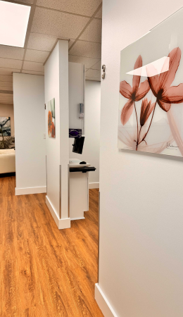 A hallway with wood flooring in a modern office or clinic, featuring floral artwork on white walls and a computer workstation in the background.