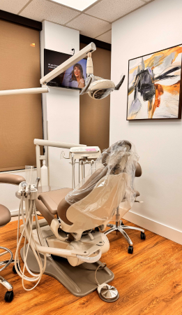 A dental exam chair with protective covering sits in a modern office. Dental equipment, abstract wall art, and a TV on the wall are visible.