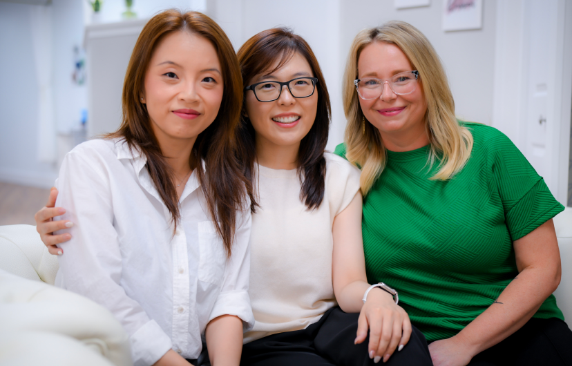 Three women are sitting together on a couch, smiling at the camera in a brightly lit indoor setting.