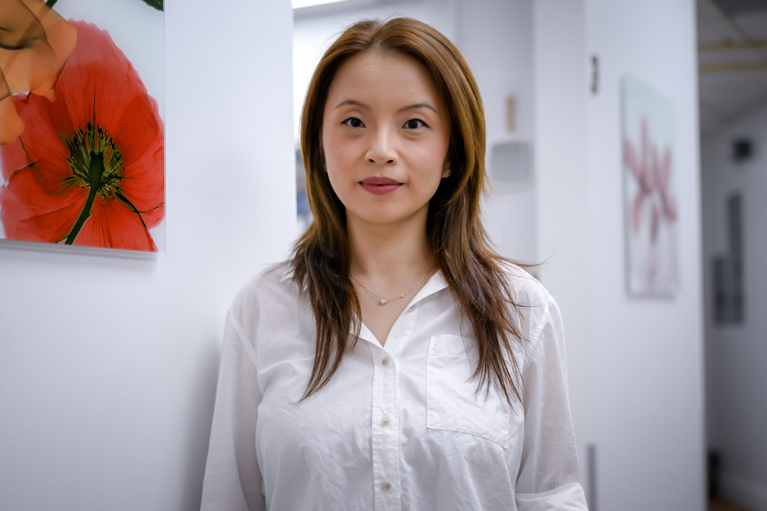 A woman with straight brown hair wearing a white button-up shirt stands indoors, with floral artwork visible on white walls behind her.