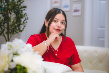 A woman in a red shirt sits on a white couch, resting her chin on her hand, with white flowers and framed pictures visible in the background.