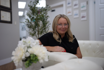 A woman with blonde hair and glasses sits on a white sofa in a modern office, with framed certificates and art on the walls, and a vase of white flowers on the table.