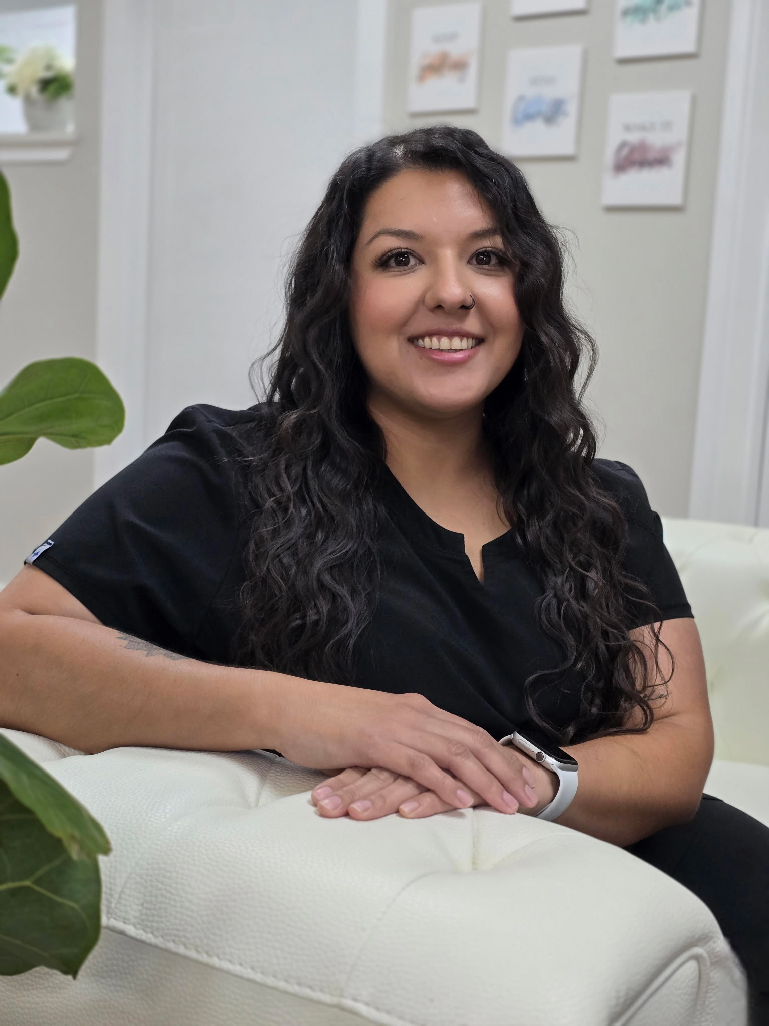 A woman with long dark hair sits on a white couch, smiling at the camera. She is wearing black clothing and a smartwatch. There are framed pictures on the wall behind her.