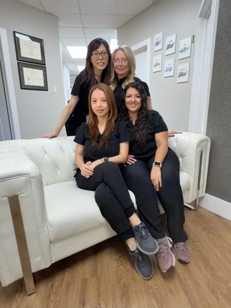 Four women in black scrubs pose together on and around a white couch in a hallway with certificates and artwork on the walls.